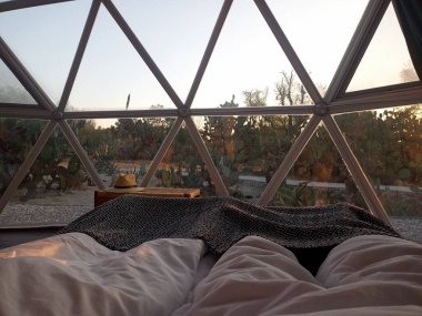 Çift yatağında uzanmış güneşin doğuşunu jeodezik çadır penceresinden izliyor.