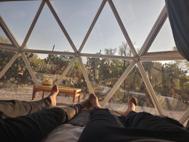 Çift yatağında uzanmış güneşin doğuşunu jeodezik çadır penceresinden izliyor.