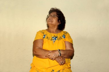 Sarı elbiseli ve gözlüklü yaşlı yerli kadın sakince oturuyor.