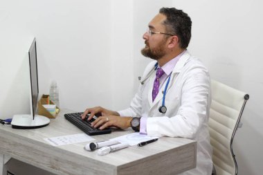 Gecelik, steteskop ve gözlüklü uzman doktor ofisinde masasında oturmuş bilgisayarla çalışıyor.