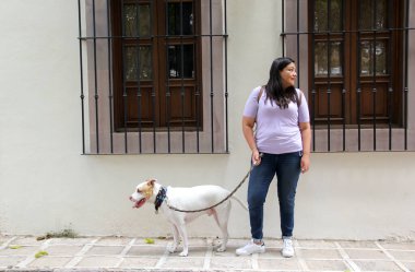 Latin yetişkin bir kadın beyaz pitbull köpeği eşliğinde koloni tarzı pencerelerle kaldırım taşından bir caddede yürüyor.