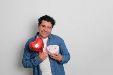 40 yaşındaki siyah saçlı ve kıvırcık saçlı Latin bir adam Sevgililer Günü 'nü kutluyor ve aşkını kalp şeklinde bir balon, gözlük, hediye ve oyuncak ayıyla gösteriyor.