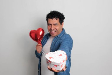 40 yaşındaki siyah saçlı ve kıvırcık saçlı Latin bir adam Sevgililer Günü 'nü kutluyor ve aşkını kalp şeklinde bir balon, gözlük, hediye ve oyuncak ayıyla gösteriyor.