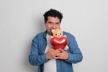 40 yaşındaki siyah saçlı ve kıvırcık saçlı Latin bir adam Sevgililer Günü 'nü kutluyor ve aşkını kalp şeklinde bir balon, gözlük, hediye ve oyuncak ayıyla gösteriyor.