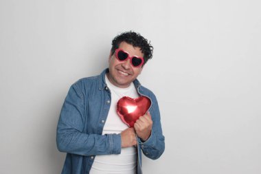 40 yaşındaki siyah saçlı ve kıvırcık saçlı Latin bir adam Sevgililer Günü 'nü kutluyor ve aşkını kalp şeklinde bir balon, gözlük, hediye ve oyuncak ayıyla gösteriyor.