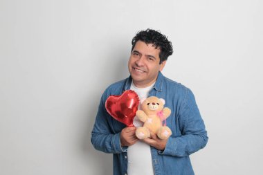 40 yaşındaki siyah saçlı ve kıvırcık saçlı Latin bir adam Sevgililer Günü 'nü kutluyor ve aşkını kalp şeklinde bir balon, gözlük, hediye ve oyuncak ayıyla gösteriyor.