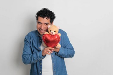 40 yaşındaki siyah saçlı ve kıvırcık saçlı Latin bir adam Sevgililer Günü 'nü kutluyor ve aşkını kalp şeklinde bir balon, gözlük, hediye ve oyuncak ayıyla gösteriyor.