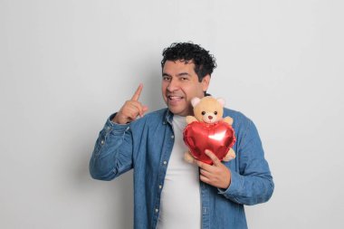 40 yaşındaki siyah saçlı ve kıvırcık saçlı Latin bir adam Sevgililer Günü 'nü kutluyor ve aşkını kalp şeklinde bir balon, gözlük, hediye ve oyuncak ayıyla gösteriyor.