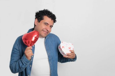 40 yaşındaki siyah saçlı ve kıvırcık saçlı Latin bir adam Sevgililer Günü 'nü kutluyor ve aşkını kalp şeklinde bir balon, gözlük, hediye ve oyuncak ayıyla gösteriyor.