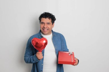 40 yaşındaki siyah saçlı ve kıvırcık saçlı Latin bir adam Sevgililer Günü 'nü kutluyor ve aşkını kalp şeklinde bir balon, gözlük, hediye ve oyuncak ayıyla gösteriyor.