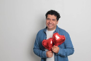 40 yaşındaki siyah saçlı ve kıvırcık saçlı Latin bir adam Sevgililer Günü 'nü kutluyor ve aşkını kalp şeklinde bir balon, gözlük, hediye ve oyuncak ayıyla gösteriyor.