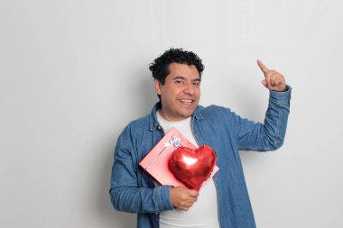 40 yaşındaki siyah saçlı ve kıvırcık saçlı Latin bir adam Sevgililer Günü 'nü kutluyor ve aşkını kalp şeklinde bir balon, gözlük, hediye ve oyuncak ayıyla gösteriyor.