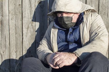Evsiz. Paçavra elbiseli ve tıbbi maskeli bir adam ahşap bir çitin yanında oturuyor. COVID-19. Kapat..