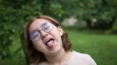 A teenage girl with glasses makes funny faces at the camera in the park. Foolishness and fun.