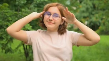Gözlüklü, kızıl saçlı tatlı bir genç kız yazın parkta gülüyor ve kamerada oynaşıyor. Komik kız. 4k video biçimi