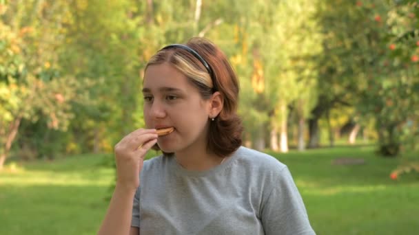 緑の自然を背景に10代の女の子がクッキーを食べます チューイン迅速かつ執拗に — ストック動画
