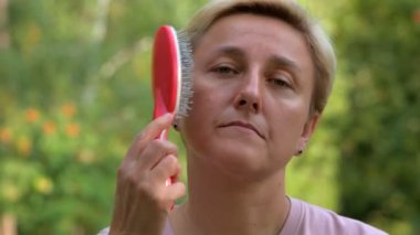 Kısa saçlı sarışın bir kız saçını yeşil arka planda kırmızı bir tarakla tarıyor.