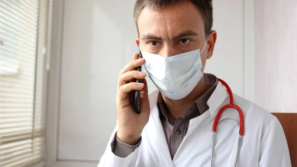 El médico preocupado con una máscara quirúrgica en la cara está hablando por teléfono —  Fotos de Stock