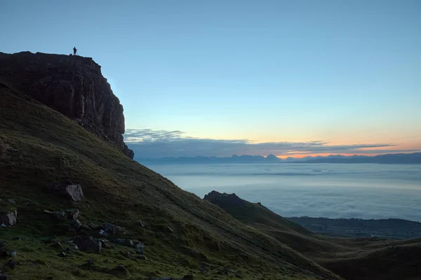 Büyük bir kayanın üzerinde duran ve güneş doğmadan önce denize bakan bir gezginin silueti.