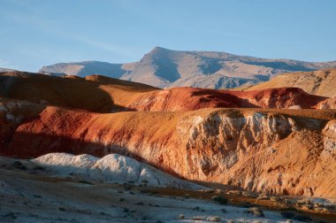 Altai Dağları 'ndaki renkli kırmızı ve turuncu tepelerin göz kamaştırıcı manzarası, sıcak akşam güneşiyle aydınlanıyor. Katmanlı dokuları ve açık mavi gökyüzü olan doğal manzara..