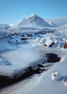 Glencoe 'da ikonik Buachaille Etive Mor' un Etive Nehri 'nin üzerinde yükseldiği parlak, dondurucu kış sahnesi. Donmuş nehir ve karla kaplı kayalar açık mavi gökyüzünün altında ön plana hakim.