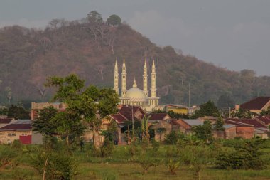Endonezya 'nın kırsal kesimlerinde ve yeşil yaprakların arasında yer alan, birden fazla minare ve merkezi kubbeden oluşan, soluk sarı bir cami yapısının manzarası, bulutlu bir gökyüzünün altında uzanan kuru, ormanlık bir tepe..