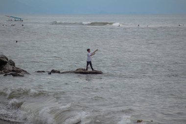 LOMBOK, İNDONEZYA - 16 ARALIK, 2025 - Gelgit kayasının üzerinde duran yalnız bir erkek balıkçının dramatik bulutlu bir gökyüzü altında çizgilerini dalgalı deniz suyuna düşürmesi.