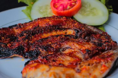Lezzetli Endonezya Ikan Bakar 'ın iştah açıcı ayrıntıları keskin kırmızı biber baharatında marine edilmiş ızgara balık geleneksel olarak salatalık ve domates dilimleriyle süslenmiş beyaz bir tabakta servis edilir..