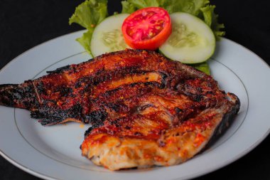 Lezzetli ve aromatik Ikan Bakar (Endonezya Izgara Balığı) tatlı ve baharatlı sambal ile kaplı, beyaz bir tabakta marul, domates ve salatalık dilimleri ile koyu bir zemin üzerinde profesyonel olarak sunulur..