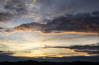 Karanlık bulutların silueti ve ufukta parlayan altın ışıkla birlikte dramatik günbatımı gökyüzü, doğa arka planını oluşturuyor.