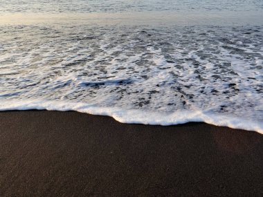 Dalgalar, doğal ışık altında köpüklü dokularla siyah kumsalda yavaşça dolaşarak rahatlama ve kıyı kaçamağı için sakin bir manzara yaratıyor.