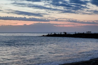 Siluetli bir grup, kayalık sahil şeridinde gün batımının keyfini çıkarıyor. Doğal ışık tarafından vurgulanıyor. Rahatlamak ve seyahat etmek için huzurlu bir sahil görüntüsü.