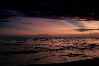 Dalgalanan Suyla Sahil Günbatımı, Kara Bulutlar ve Huzurlu bir Manzara Yaratan Doğal Işık Yansımaları