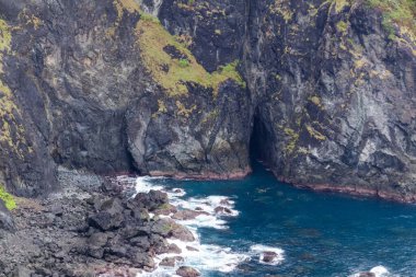 Doğal mağara girişi ve derin mavi okyanus sularının kayalık kıyı şeridine çarptığı engebeli karanlık volkanik deniz uçurumu duvarı..