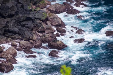 Çarpışan okyanus dalgaları koyu kıyı volkanik kayalara karşı beyaz köpük oluşturarak engebeli bir kıyı dokusu oluşturur..