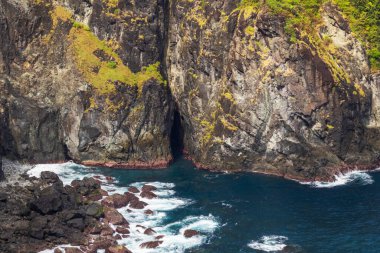Deniz mağarası girişi ve derin mavi suyu aşağıdaki pürüzlü kayalara çarpan koyu kıyı kayalıkları..