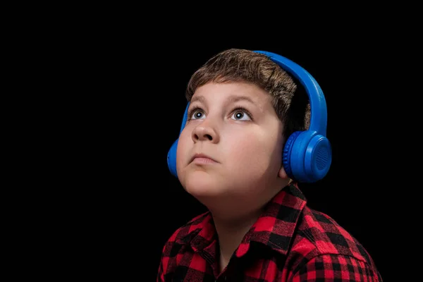 クローズアップ顔のティーン男の子で遊んだ赤いシャツで音楽を聴くで大きな青い無線耳のヘッドフォンで隔離された黒の背景 — ストック写真