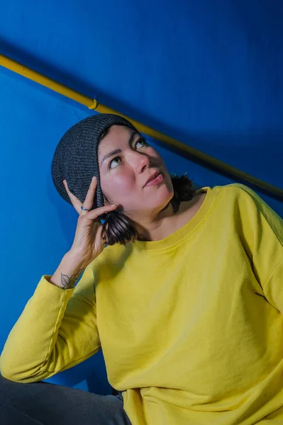 young teenage girl in a yellow sweatshirt and a knit cap props her head with her hand hand on a blue background