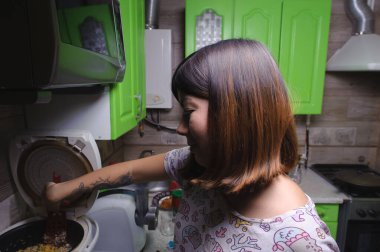 Gülümseyen, siyah saçlı, sebzeli pilav karıştıran genç bir kadın. Mutfaktaki çoklu pişirme makinesinde akşam yemeği için hazırlanmış bir spatula kullanıyor..