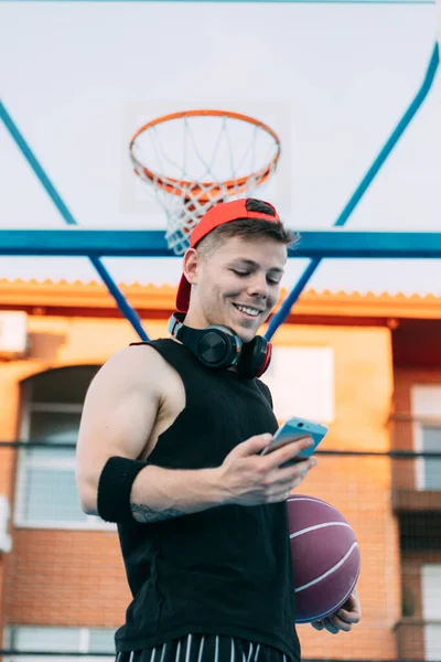 ボールを持つ魅力的な男バスケットボール選手は休憩を取りながら彼の携帯電話と笑顔を見て — ストック写真