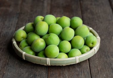 Tahta arka planda bambu kevgire yerleştirilmiş olgunlaşmamış erikler. Japon kayısı meyvesi.