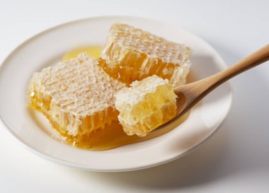 Beyaz arka planda tahtadan yapılmış kaşıkla beyaz tabakta taze organik bal peteği.