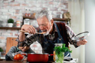 Mutlu büyükbaba ve torun mutfakta yemek pişiriyor, spatuladan insan sosu tadıyor.