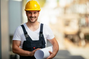 Gülümseyen inşaatçı sarı kask takıyor ve kağıt tutuyor.