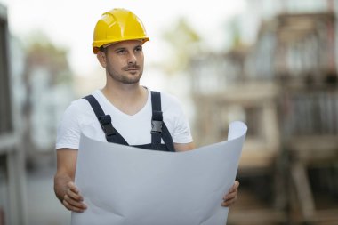 Sarı miğferli müteahhit elinde kağıt planı tutuyor ve başka tarafa bakıyor.