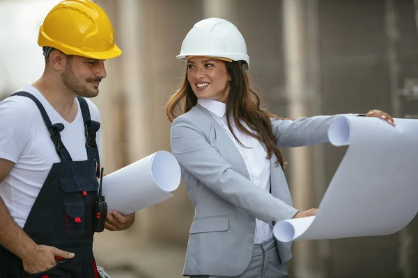 smiling man builder and woman architect in helmets holding paper plans