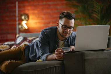 Genç adam oturma odasında online alışveriş için kredi kartı kullanıyor..