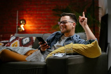 Genç adam kanepede uzanıp müzik dinliyor. Modern adam oturma odasında telefonuyla müzik dinliyor..