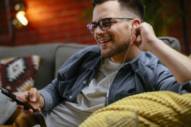 Genç adam kanepede uzanıp müzik dinliyor. Modern adam oturma odasında telefonuyla müzik dinliyor..