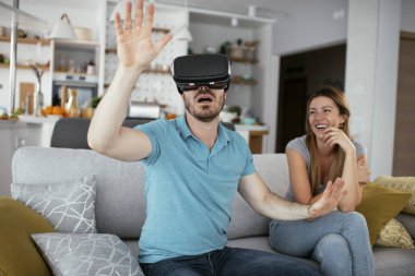 Mutlu çift oturma odasında sanal gerçeklik gözlükleriyle eğleniyor. Evde video oyunu oynayan ve eğlenen genç bir çift..